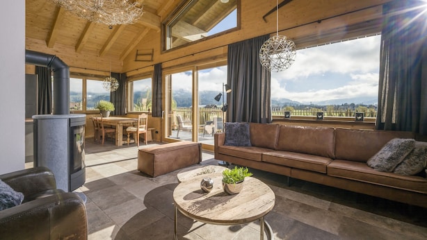 A spacious living area with a wooden ceiling, large windows, a sofa, a coffee table, and a fireplace. - Narzenhof Chalets - Familien & Luxus Apartments (Sankt Johann in Tirol)