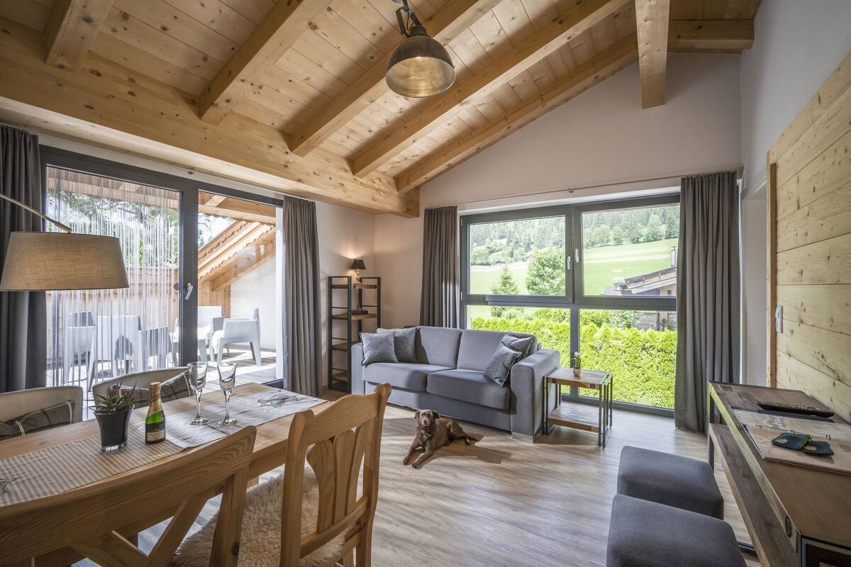 Un vaste salon doté d’un plafond en bois, d’un canapé gris, d’une table à manger avec des chaises et d’une grande fenêtre offrant une vue sur un paysage verdoyant.