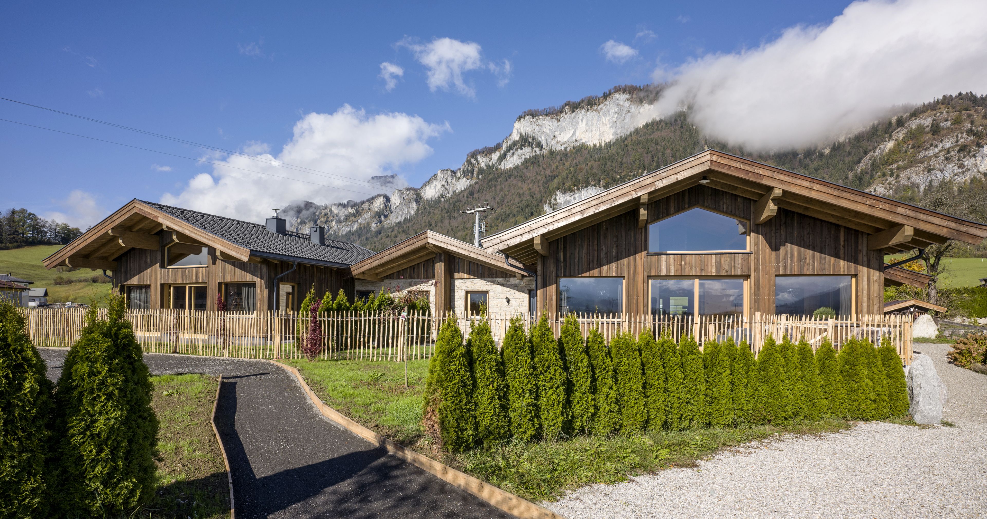 A wooden chalet with large windows, a gravel driveway, and a wooden fence.