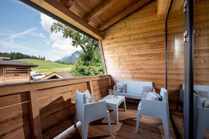 Eine Holzterrasse mit einer Couch und Stühlen, ein Tisch mit einem Glas, und Blick auf eine grüne Landschaft und Berge.