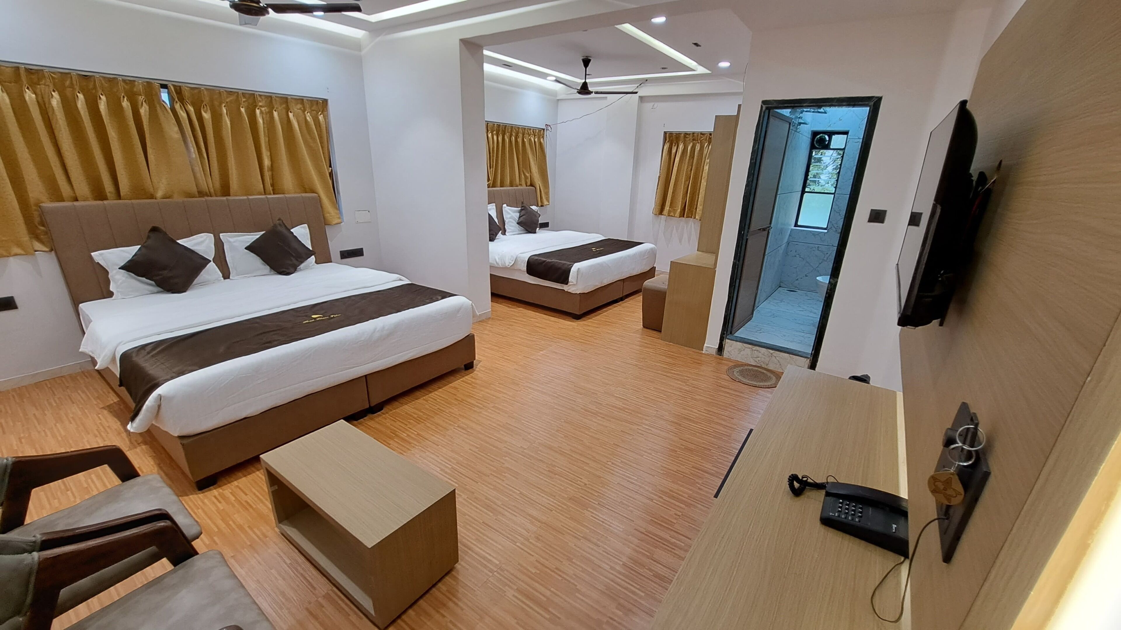 A modern hotel room with two beds, a desk, a chair, a small table, and a bathroom visible through an open door.