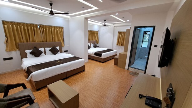 A modern hotel room with two beds, wooden flooring, ceiling fans, and a bathroom with a glass door. - Hotel Dhruv Palace (Trimbak)