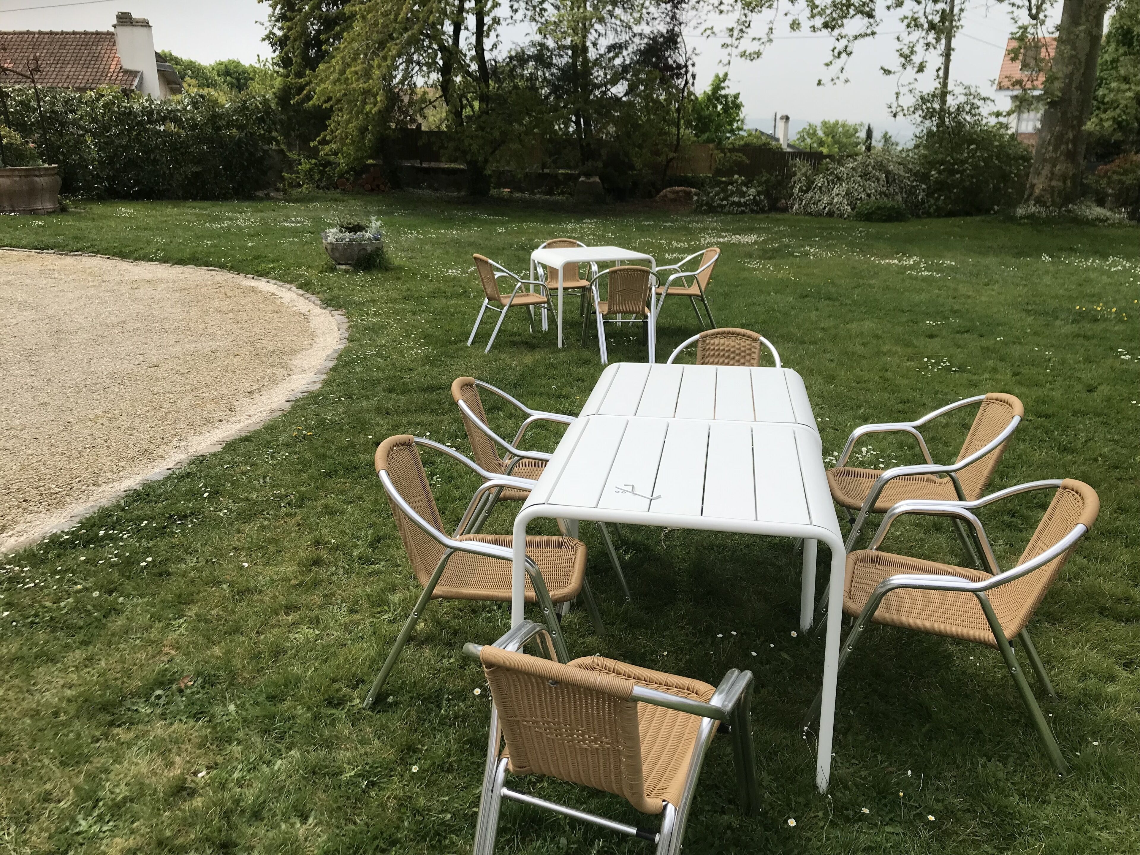 Un jardín con una mesa de metal blanco y sillas de mimbre, rodeado de césped y árboles.