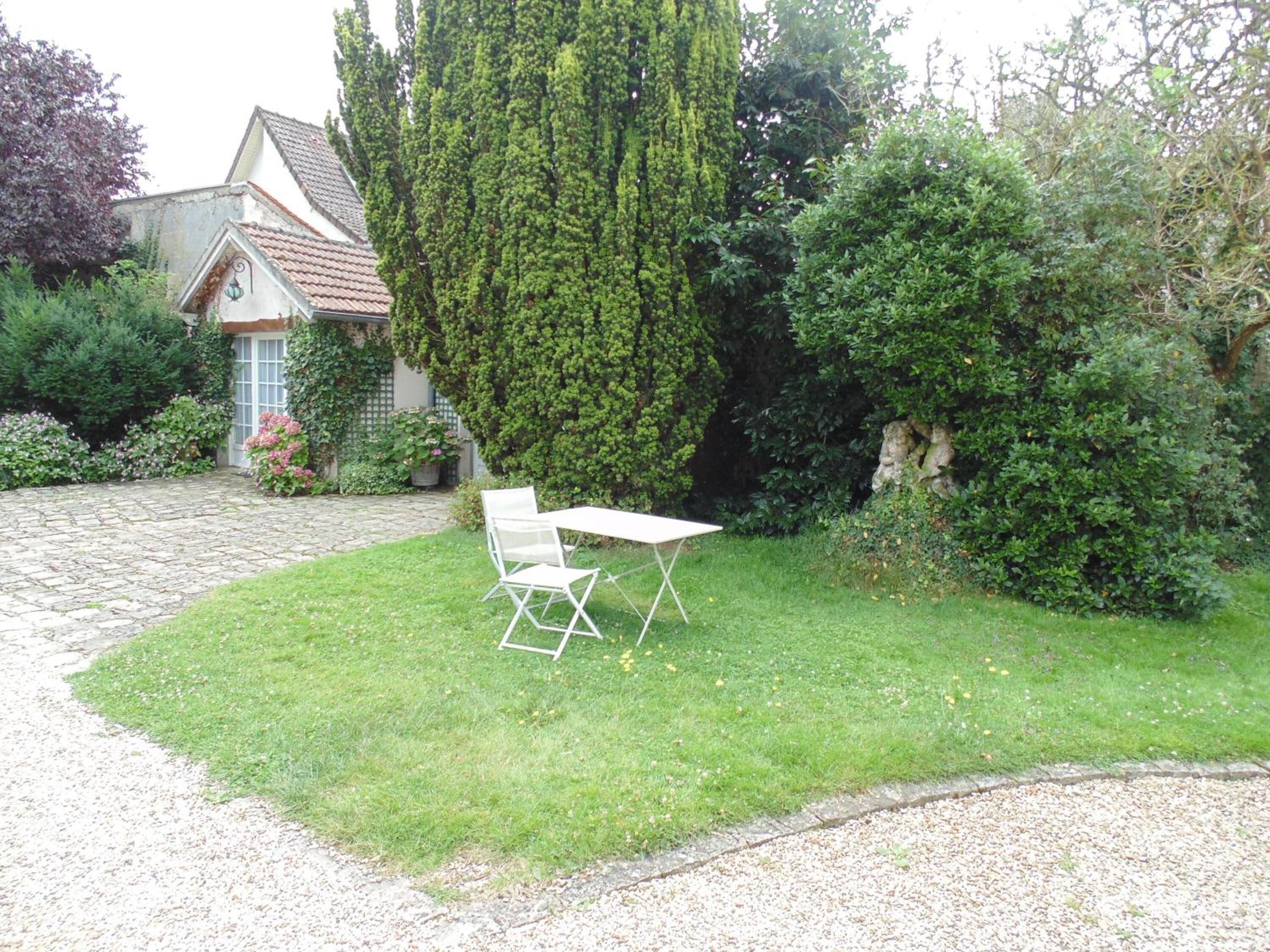 Een tuin met een witte tafel en stoelen, een kiezelpad en een huis met een pannendak.