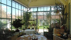 Ein Wintergarten mit rundem Tisch, gedeckt für Tee, umgeben von Topfpflanzen und Blick auf den Garten.