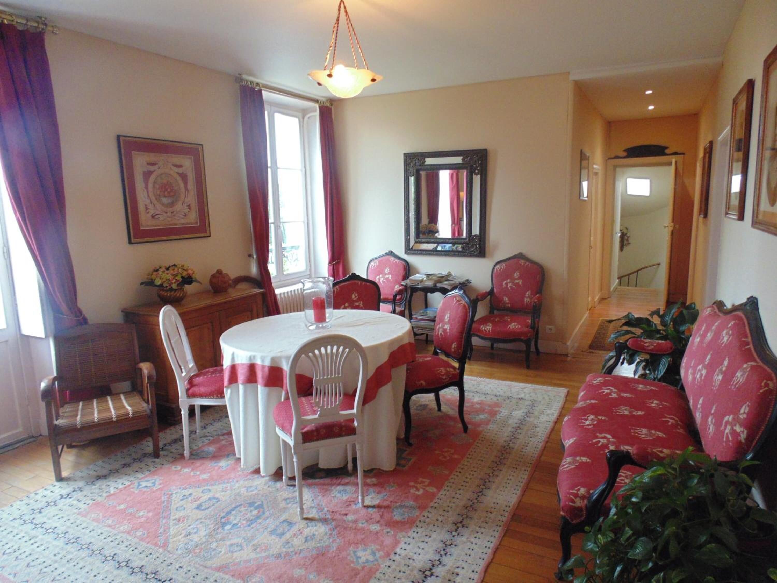 A dining area with a round table, chairs, and a red sofa. There is a mirror on the wall and a window with curtains.