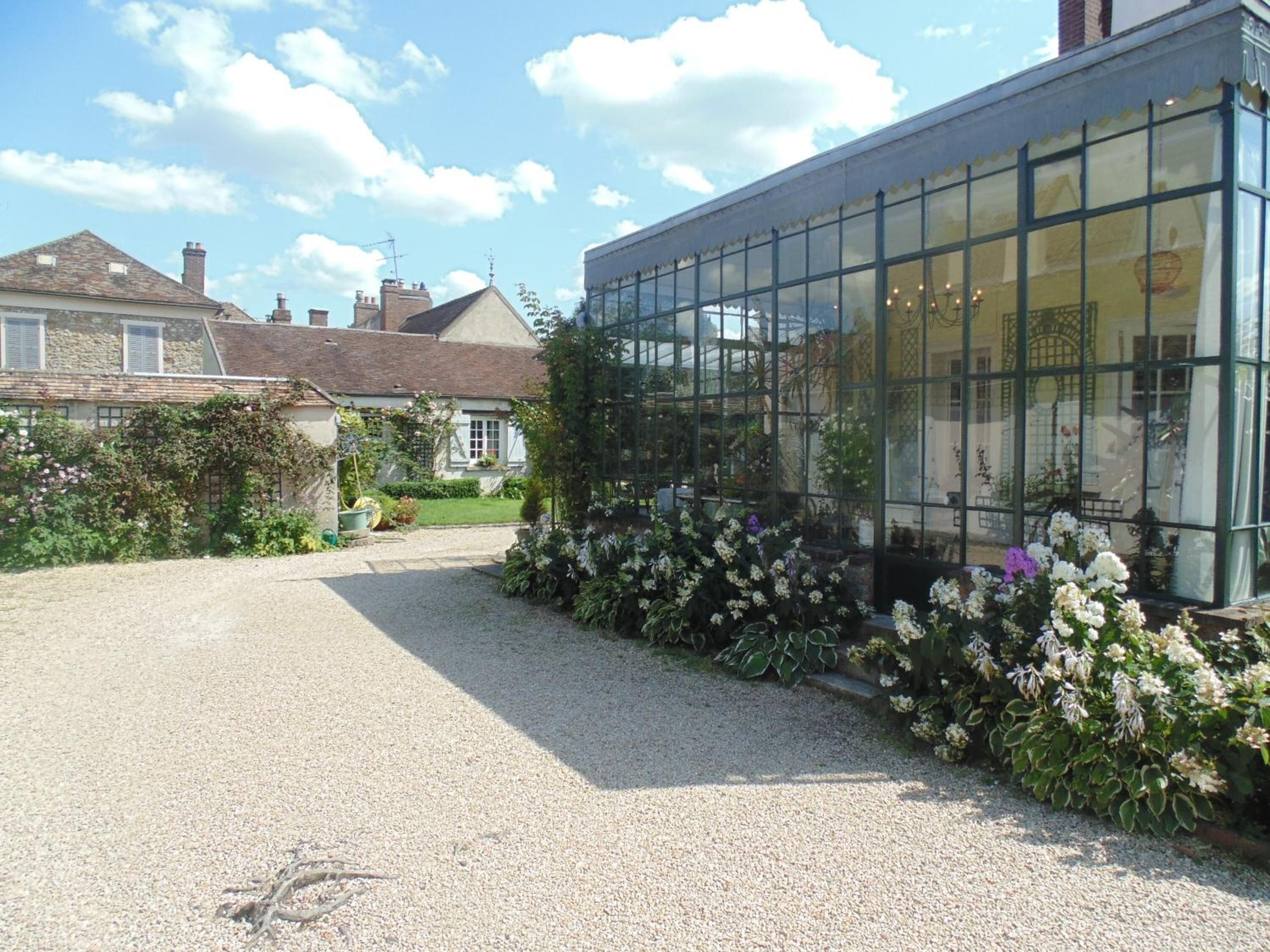 A greenhouse with a gravel driveway leading to a house with a sloped roof and chimneys.