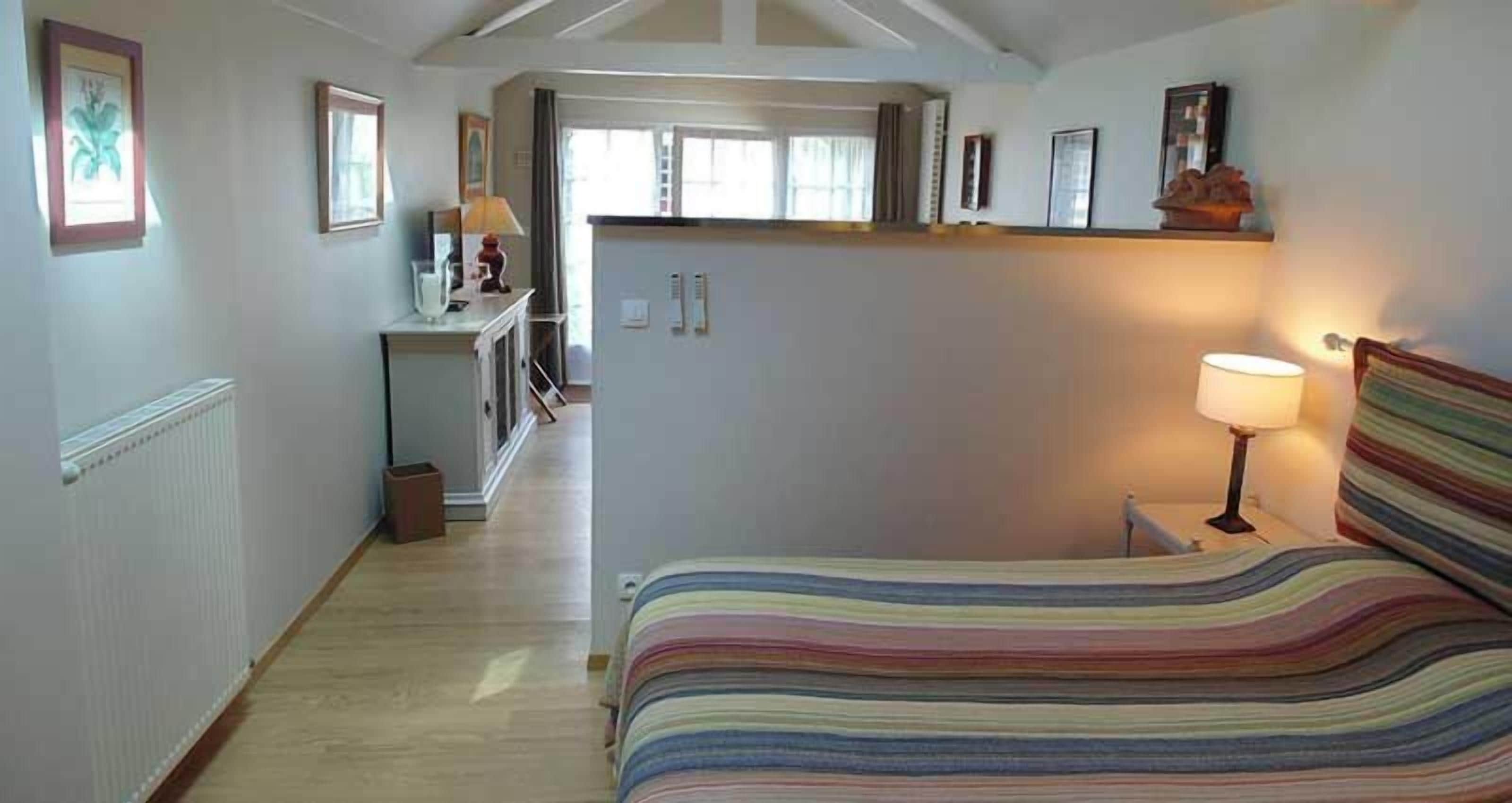 A bedroom with a bed, a nightstand, a lamp, and a view of a kitchen and dining area.