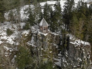 A small cabin on a snow-covered cliff surrounded by dense forest.
