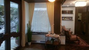 Ein gemütliches Zimmer mit Holzboden, einem Fenster mit durchscheinenden Vorhängen, einem kleinen Tisch mit Lampe und einem Sessel.