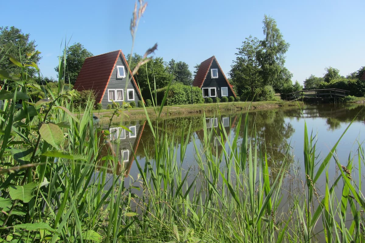 Duas casas com telhados vermelhos e molduras de janelas brancas, rodeadas por vegetação e um lago tranquilo.
