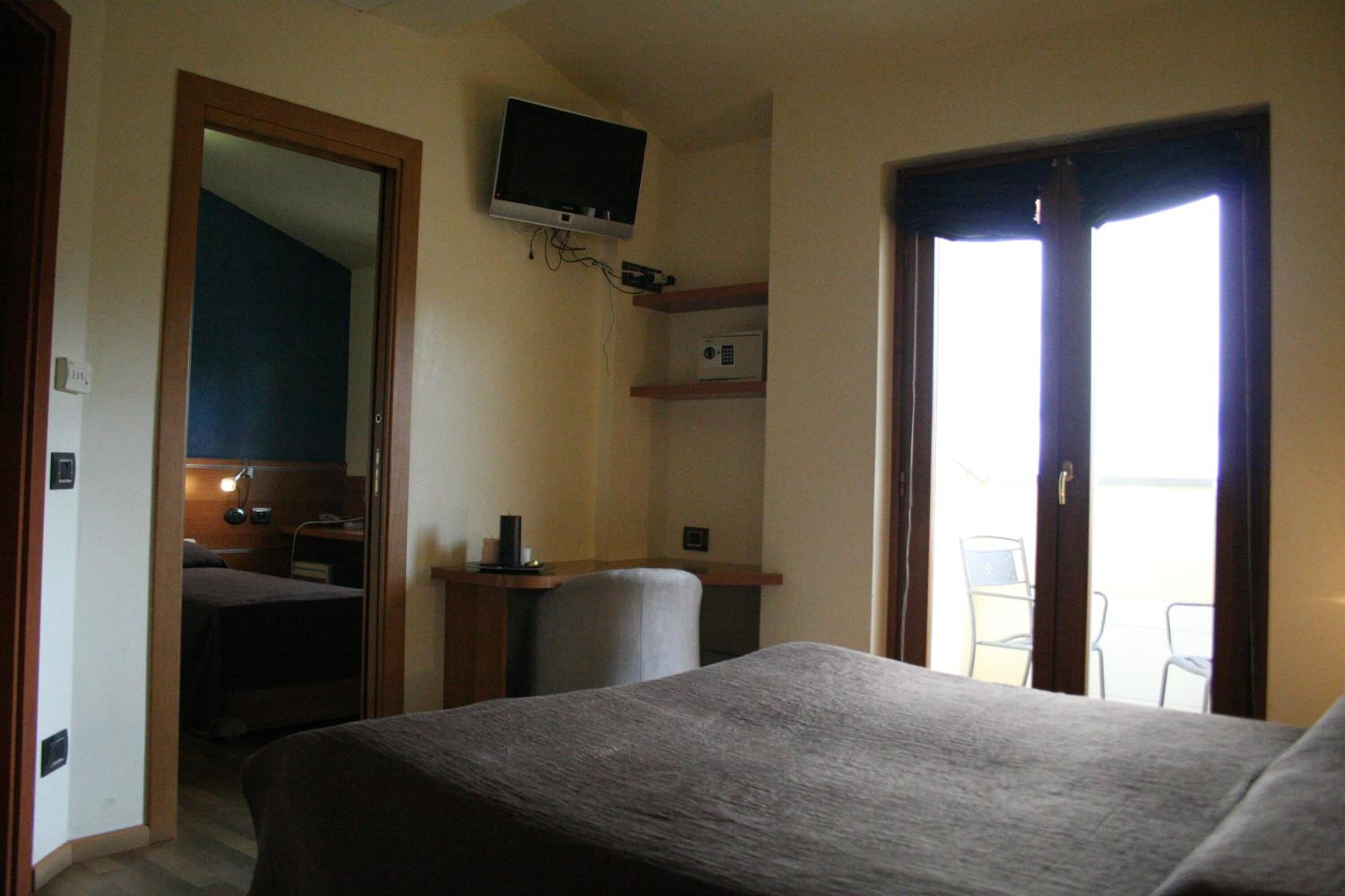 A hotel room with a bed, a desk with a chair, a television mounted on the wall, and a door leading to a balcony.