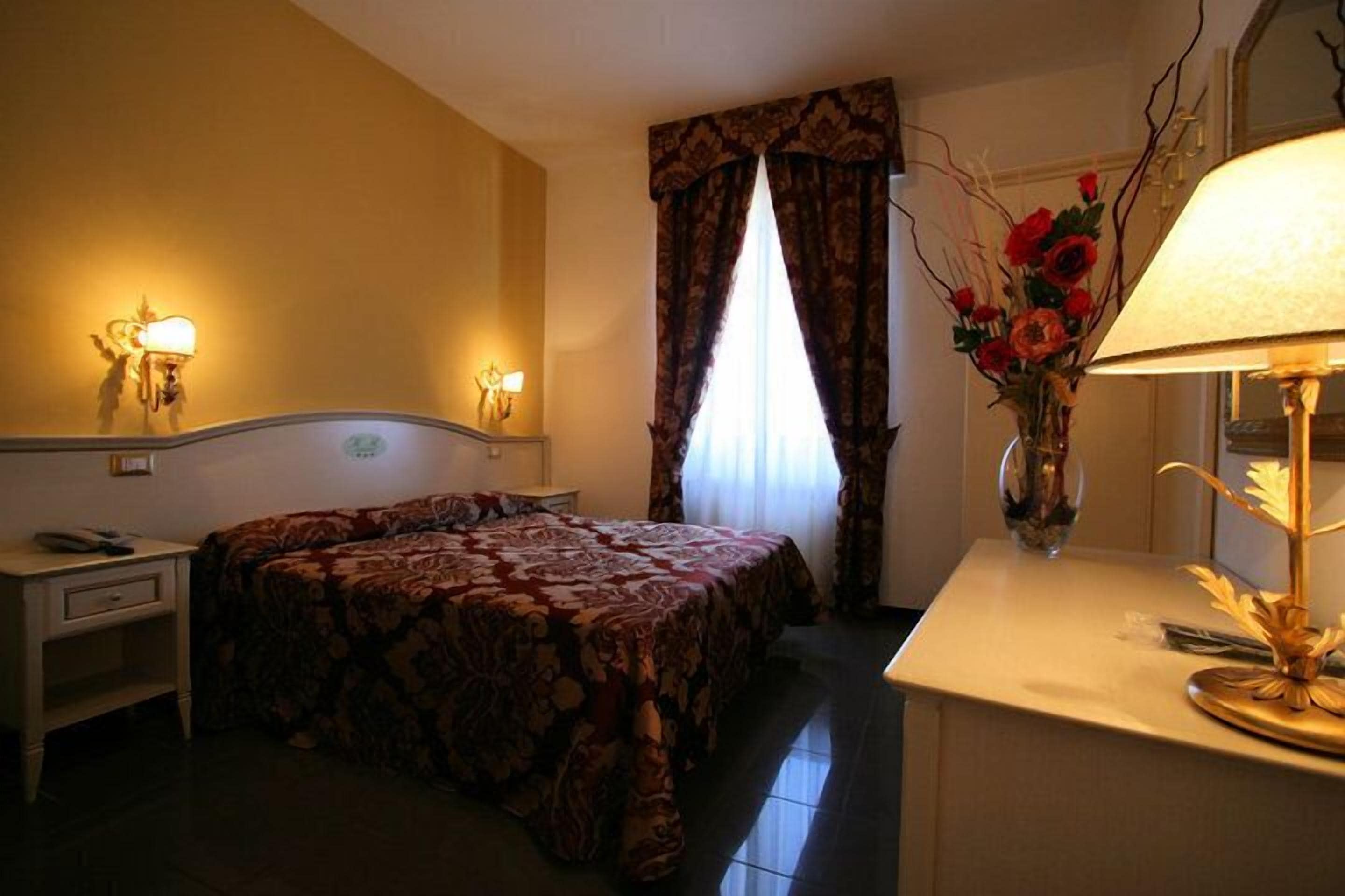 A hotel room with a bed, bedside lamps, a desk, and a vase with flowers.