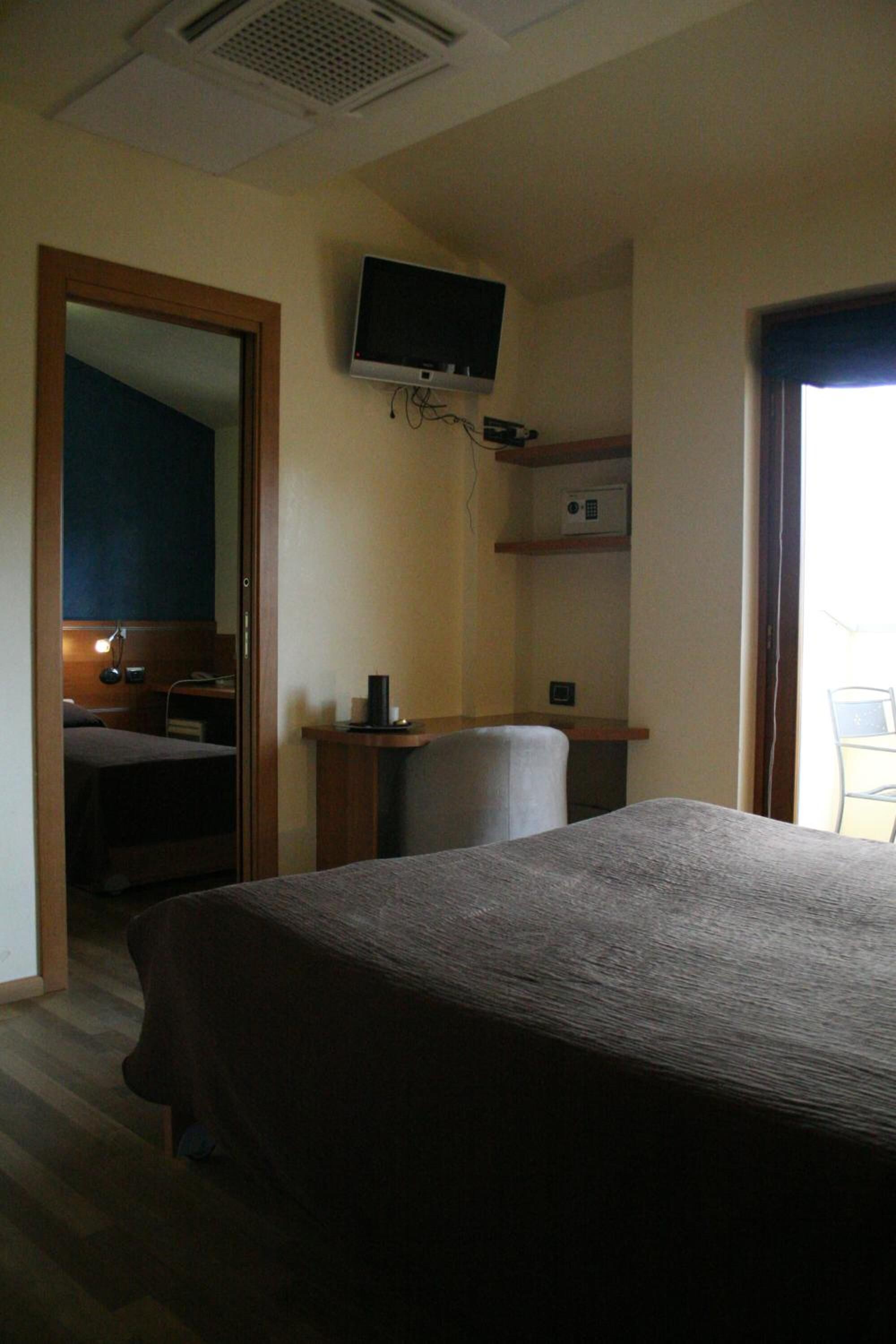 Une chambre d’hôtel équipée d’un lit, d’un bureau, d’une chaise, d’un téléviseur fixé au mur et d’une petite étagère sur laquelle se trouvent un téléphone et une tasse.