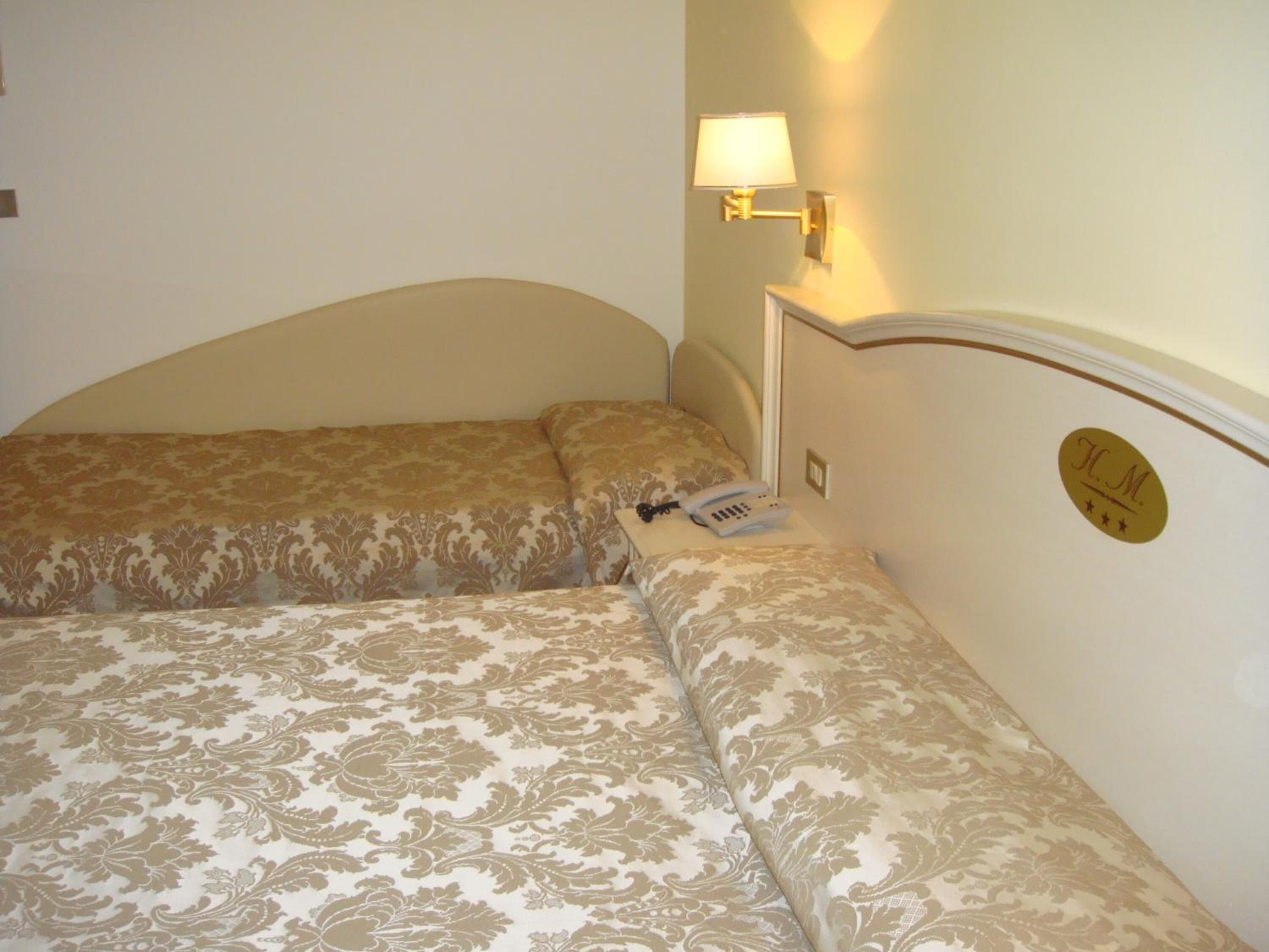 A hotel room with two beds, each with a patterned bedspread, a bedside telephone, and a wall-mounted lamp.
