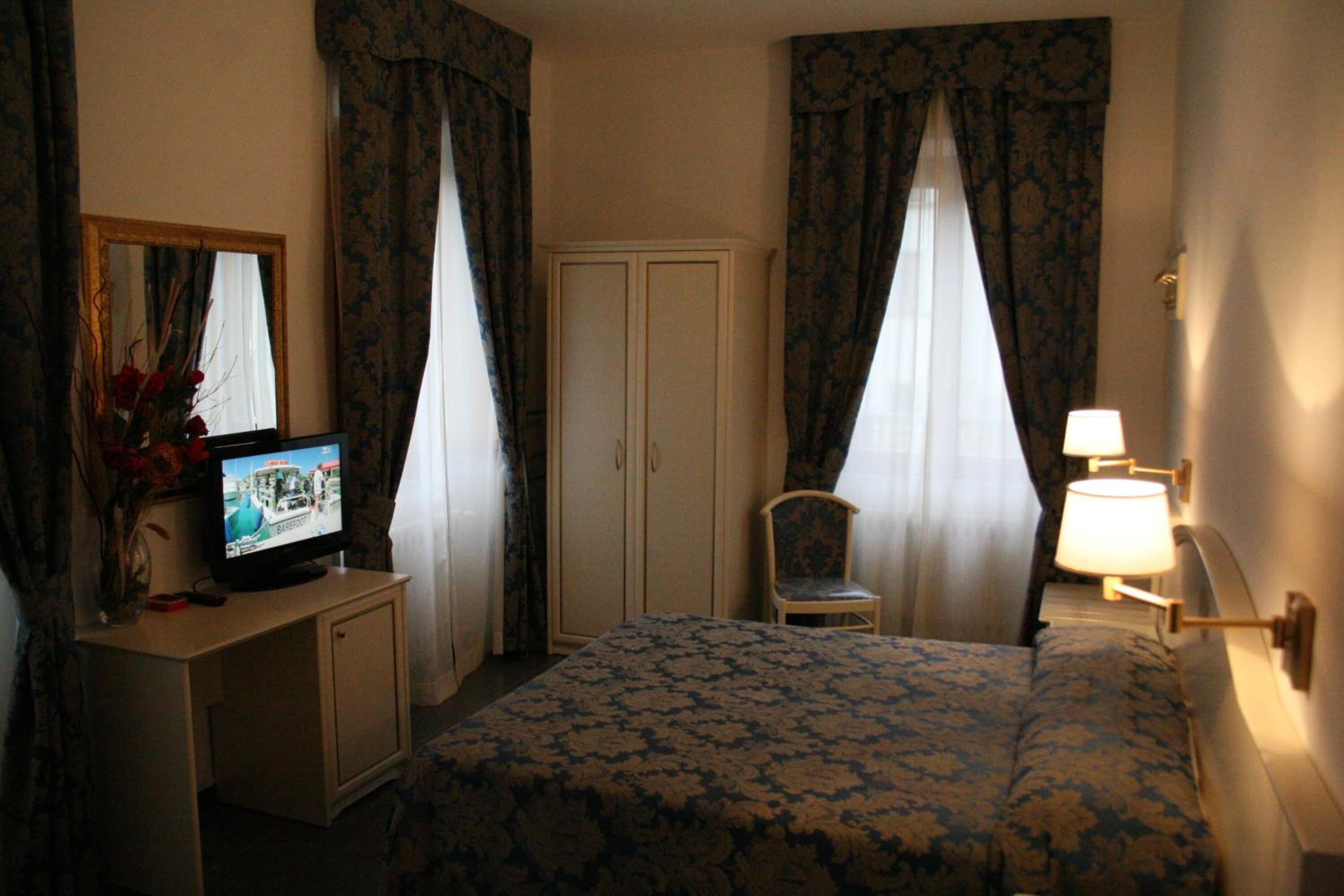A hotel room with a bed, a television on a small table, a chair, a wardrobe, and curtains.