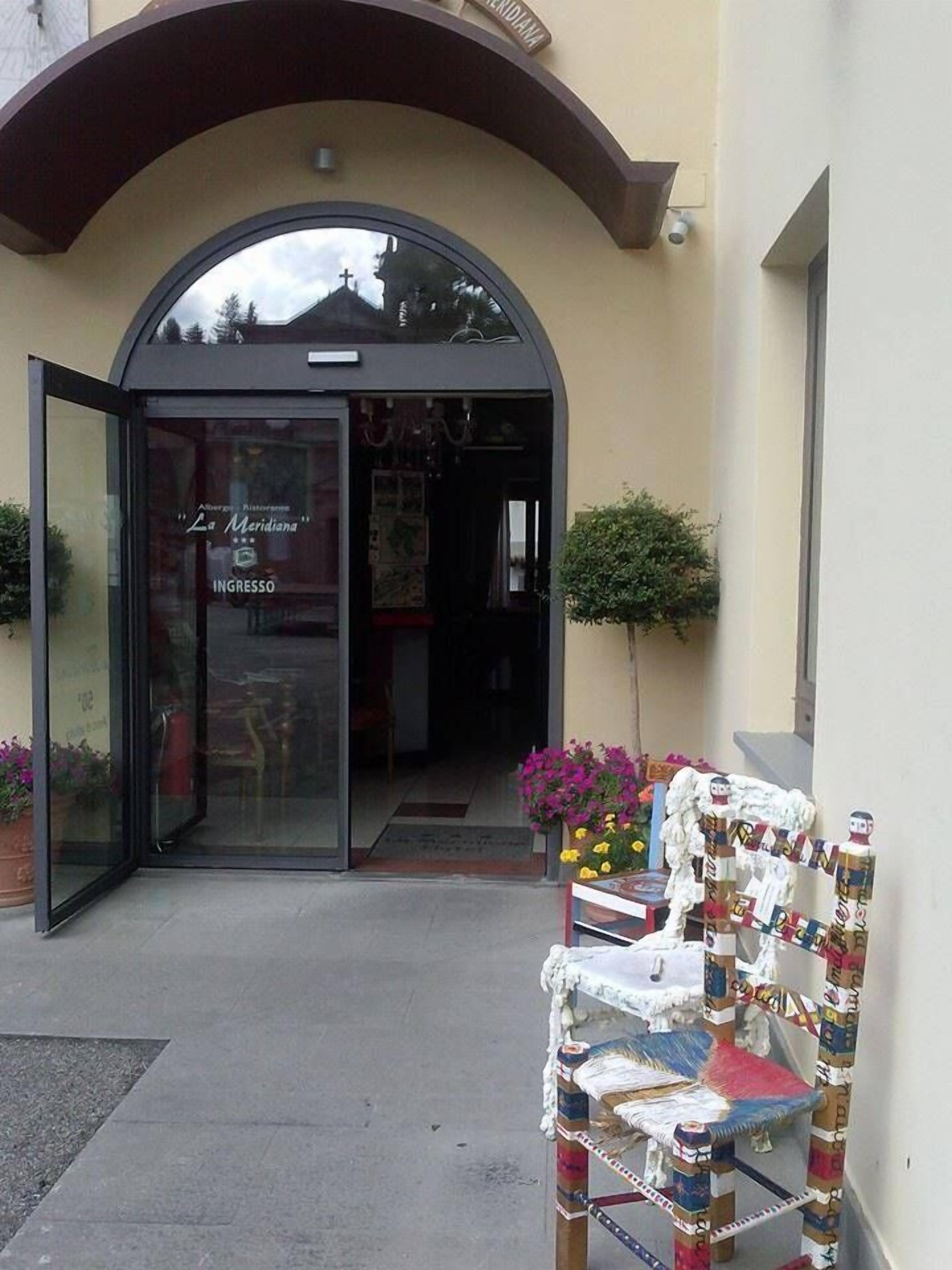 Entrada de un restaurante con puerta de cristal, un pequeño espacio exterior con una silla y plantas en macetas.