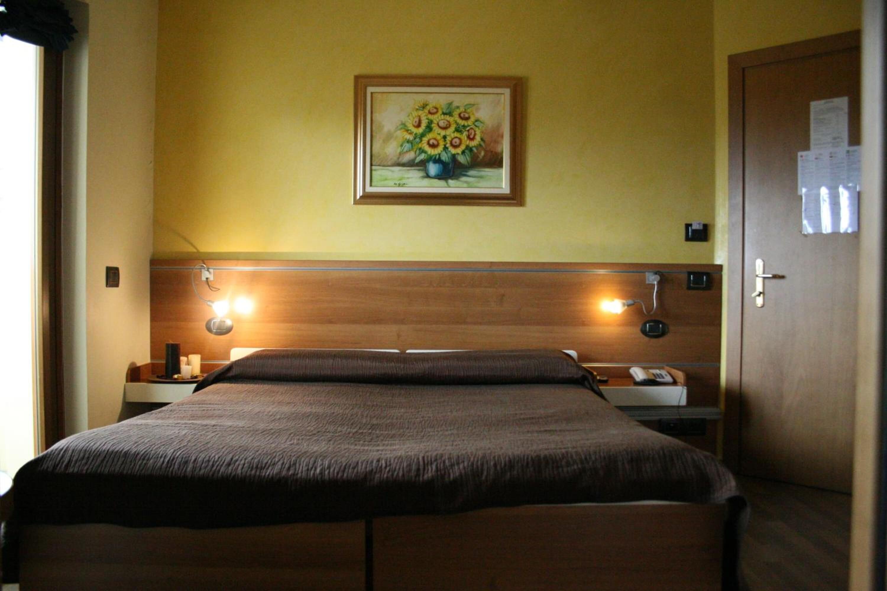 A hotel room with a bed, bedside lamps, a nightstand, a painting of sunflowers, and a door with a paper on it.