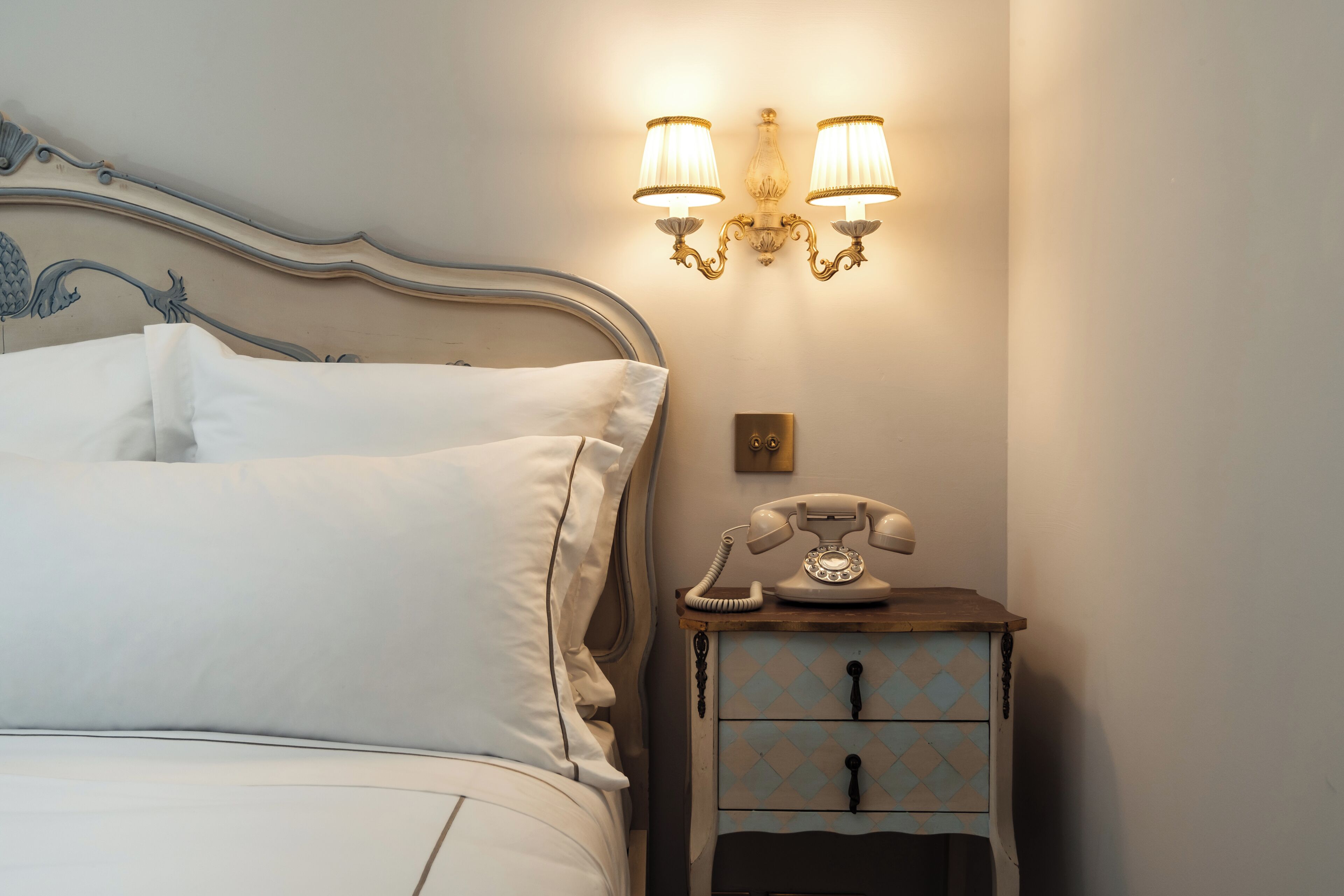 Una cama bien hecha con sábanas blancas, un teléfono de estilo vintage en la mesita de noche y una lámpara de pared.