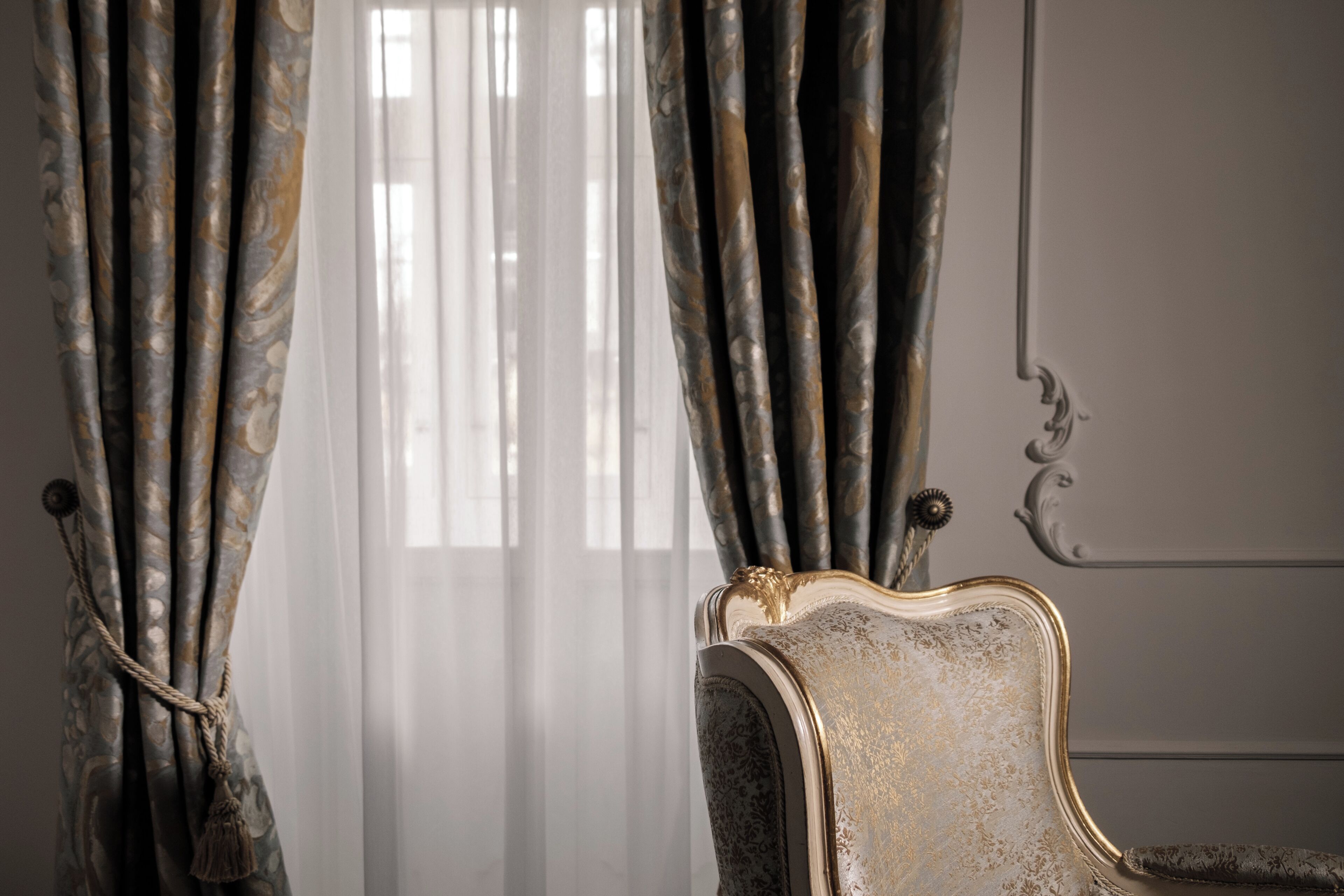 A room with a classic chair and curtains, a window with sheer curtains, and a wall with a decorative molding.