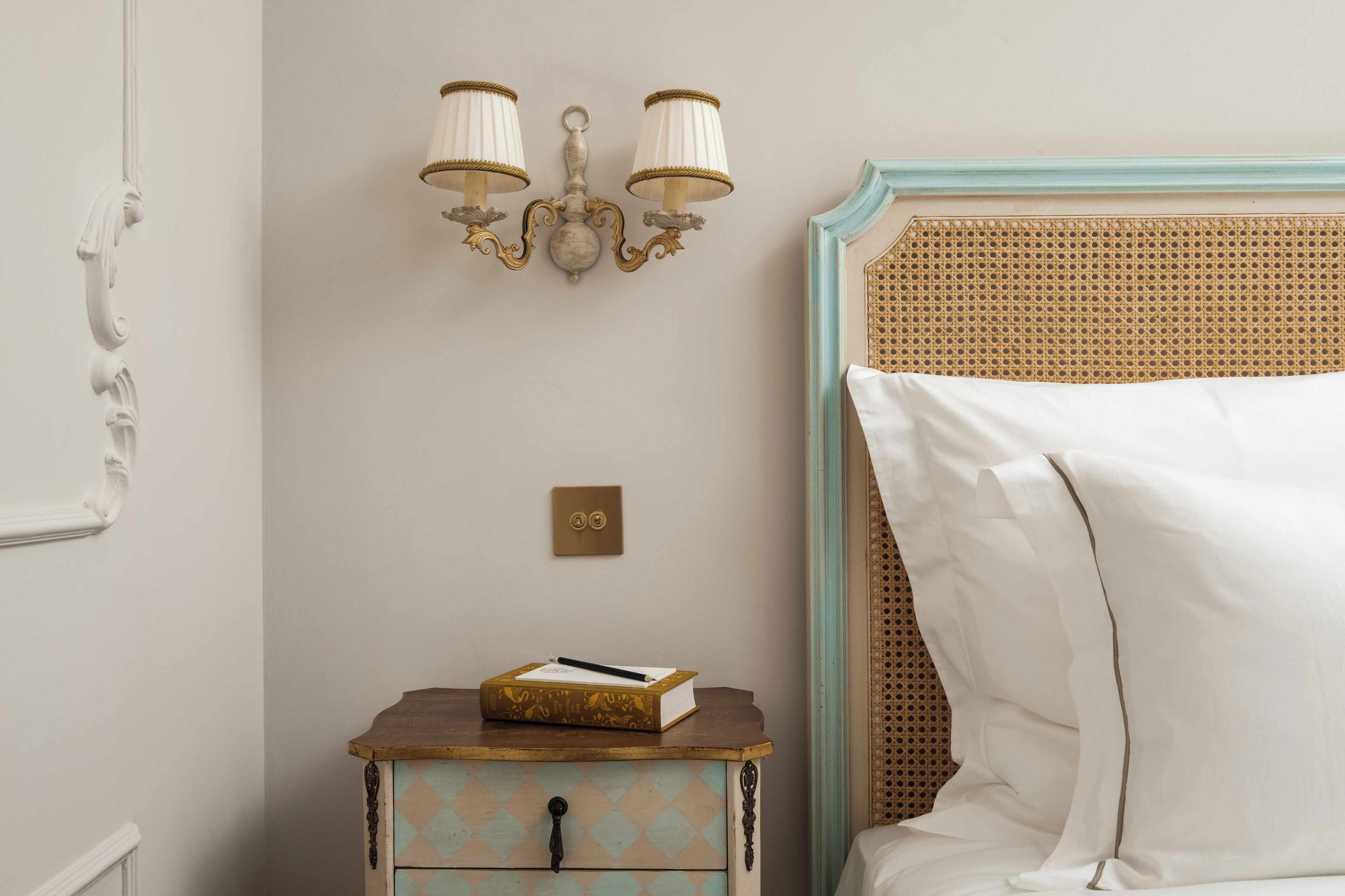 Un dormitorio con una cama, una mesita de noche con un libro y una lámpara, y una luminaria en la pared.