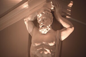 Una escultura de una figura con los brazos levantados, ubicada en una habitación con techos inclinados y vigas estructurales a la vista.