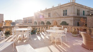 An outdoor terrace with white metal furniture, potted plants, and a large ornate building in the background. - Domus Zamittello (Valletta)
