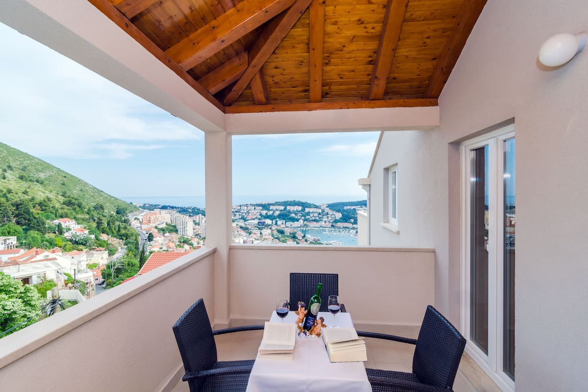 Een balkon met een houten plafond, een tafel met twee stoelen en uitzicht op een kustplaatsje.