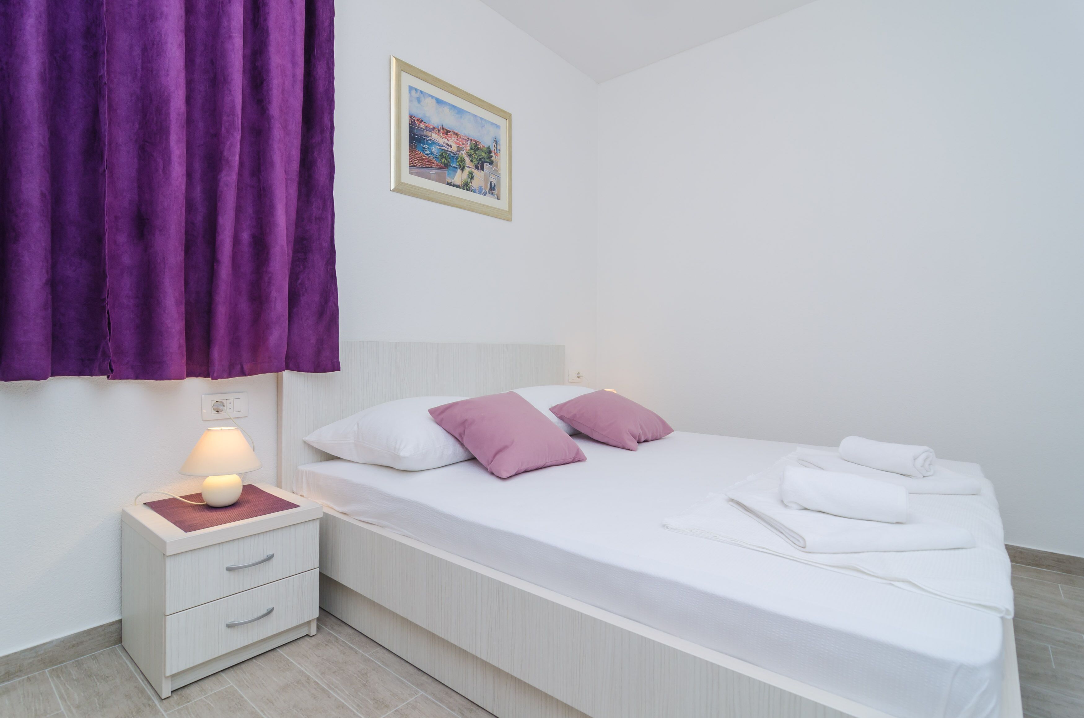 Een slaapkamer met een bed, kussens, een nachtkastje met een lamp en een raam met paarse gordijnen.