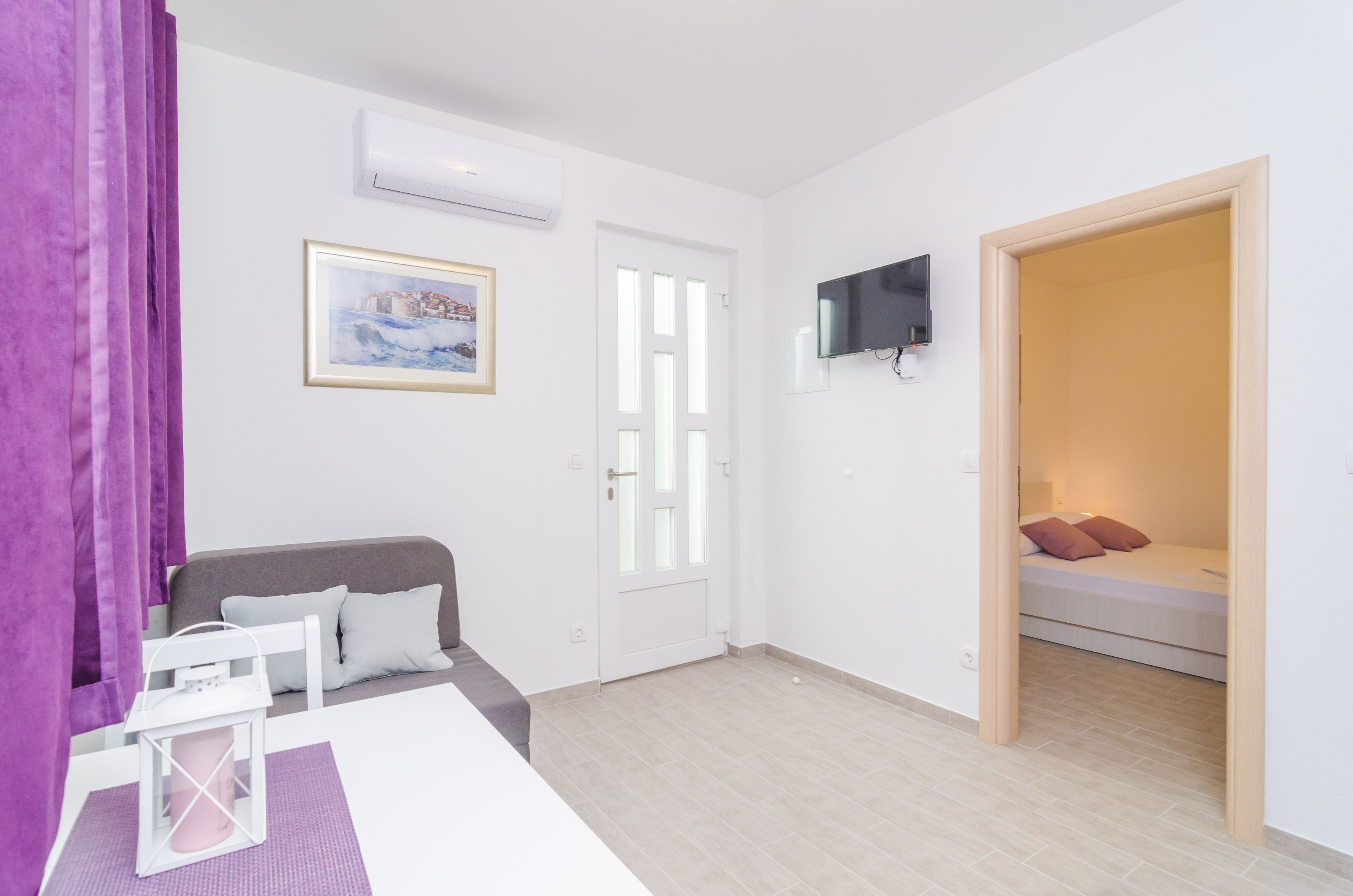 A room with a bed, a TV mounted on the wall, a framed picture, and a window with curtains.