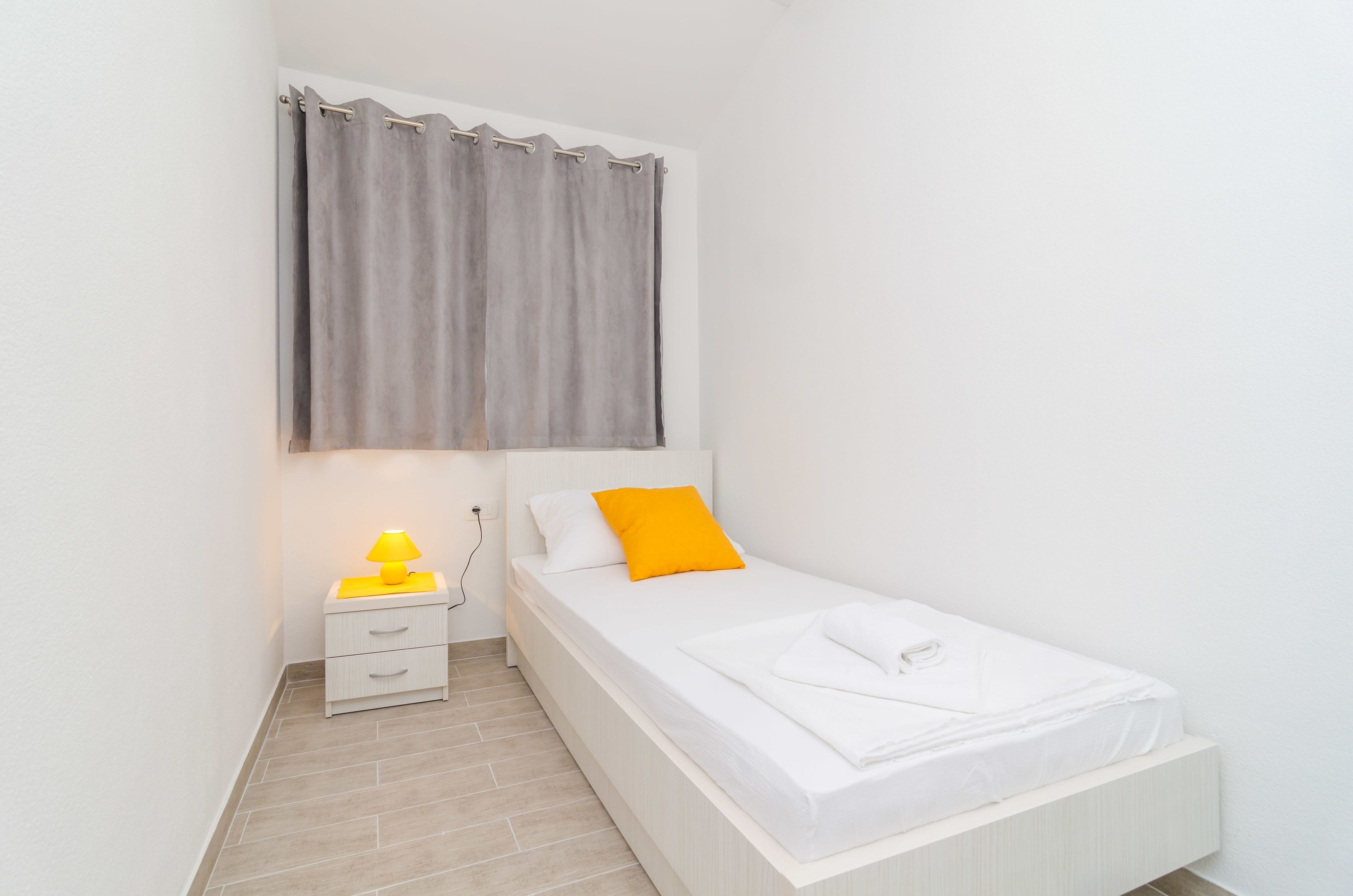 Un dormitorio pequeño y ordenado con una cama individual, una mesita de noche con lámpara y una ventana con cortinas.