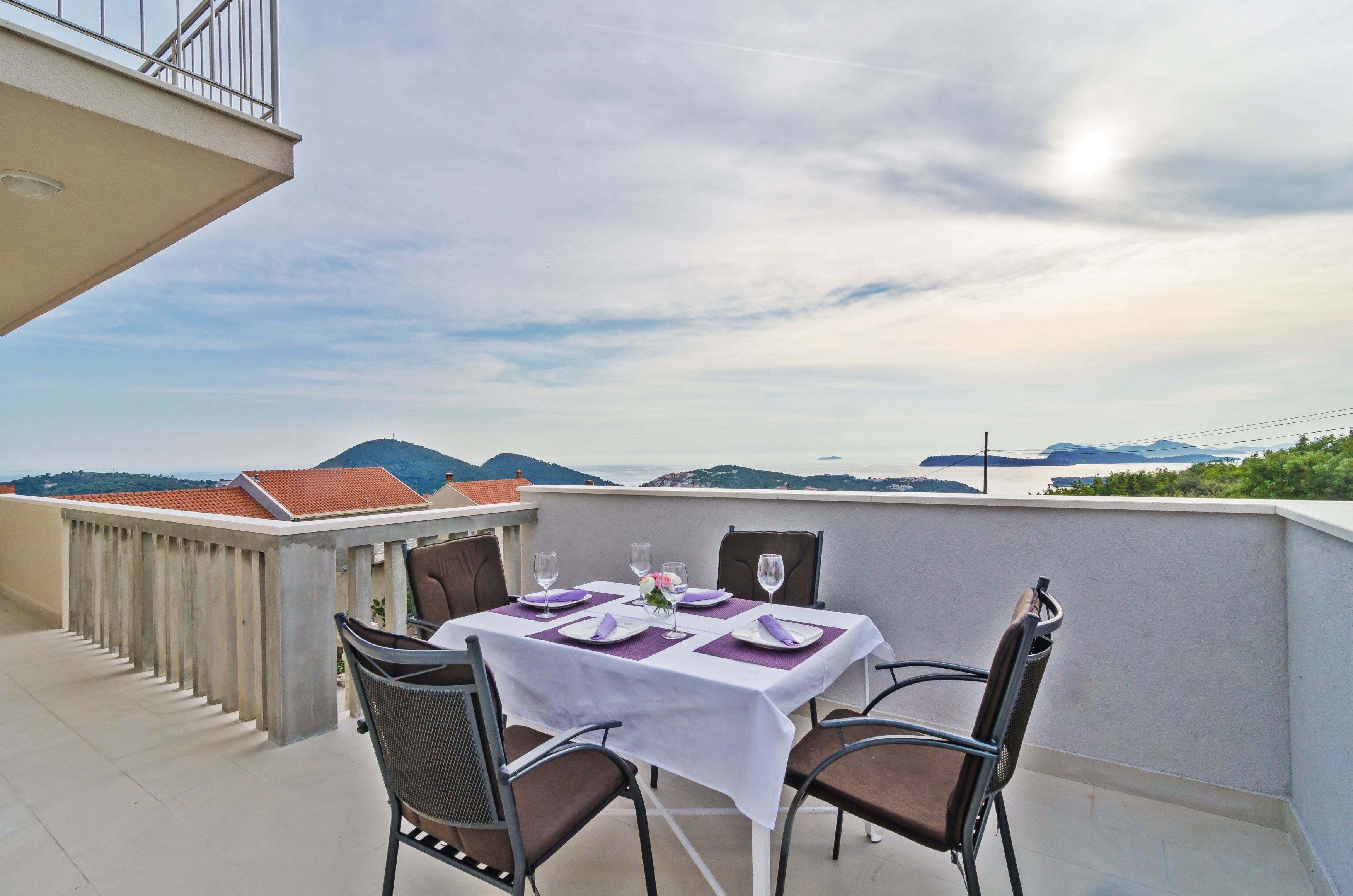 Une table dressée pour deux personnes sur un balcon offrant une vue sur les montagnes et la mer.
