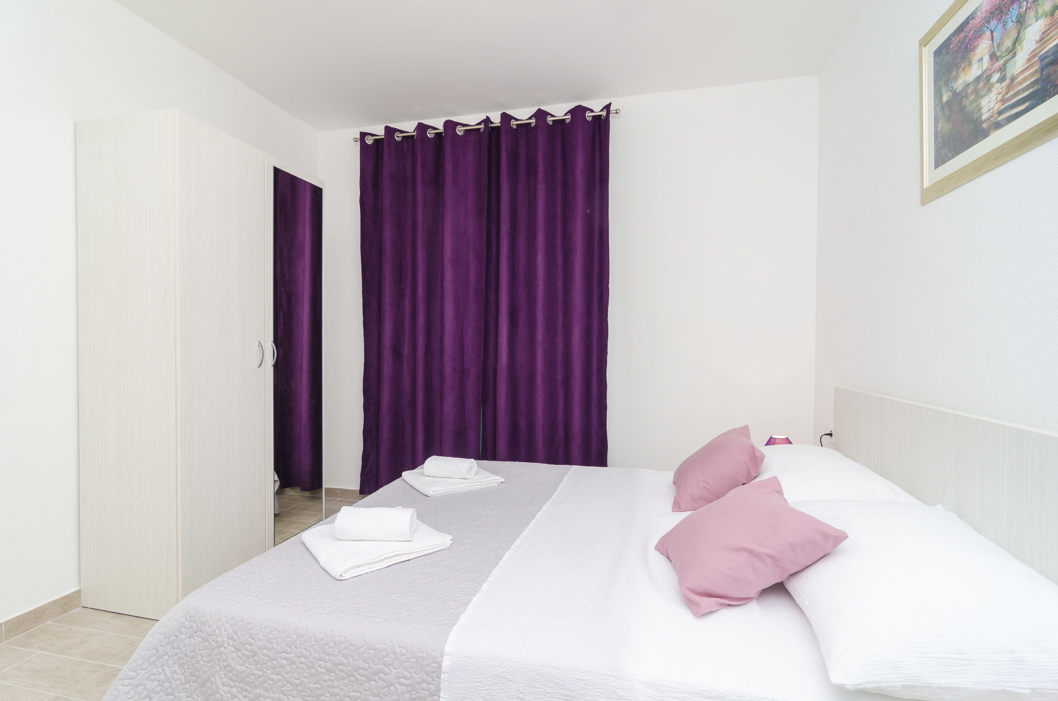 Una cama bien hecha con almohadas rosadas y blancas, una manta blanca y toallas dobladas sobre un edredón blanco.