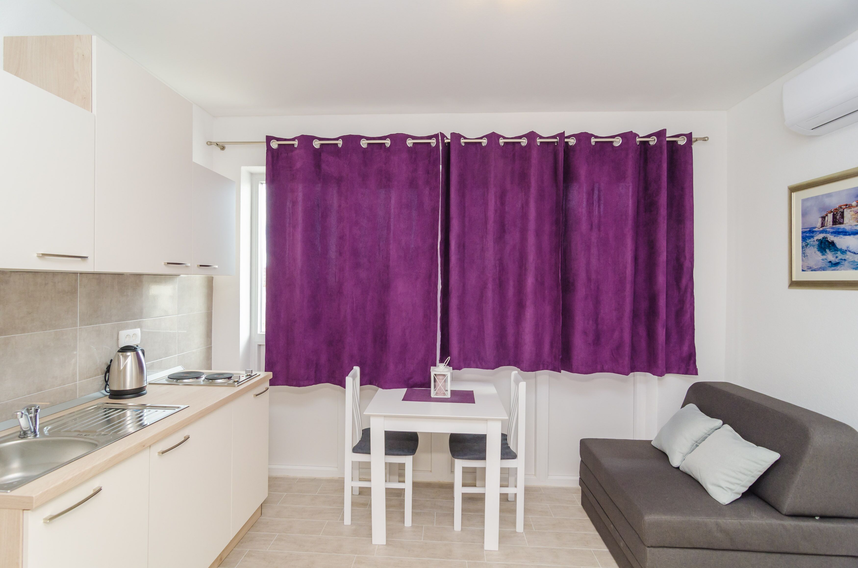 Una cocina compacta con encimera blanca, una tetera y una pequeña mesa con dos sillas. Hay un sofá gris con un cojín y una cortina morada que cubre la ventana.