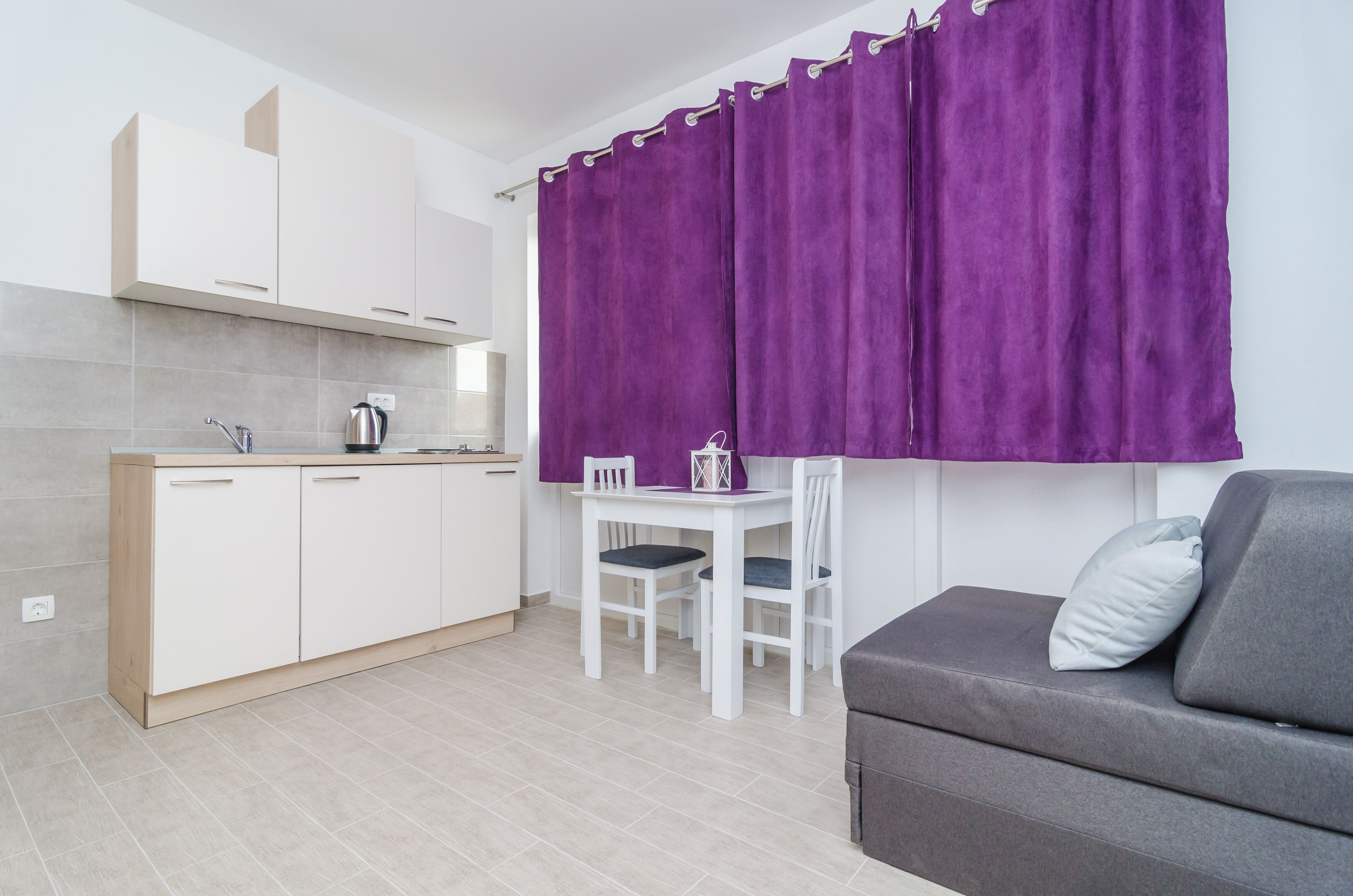 A compact kitchen with white cabinets, a dining table with two chairs, and a grey sofa with a pillow. Purple curtains cover the window.