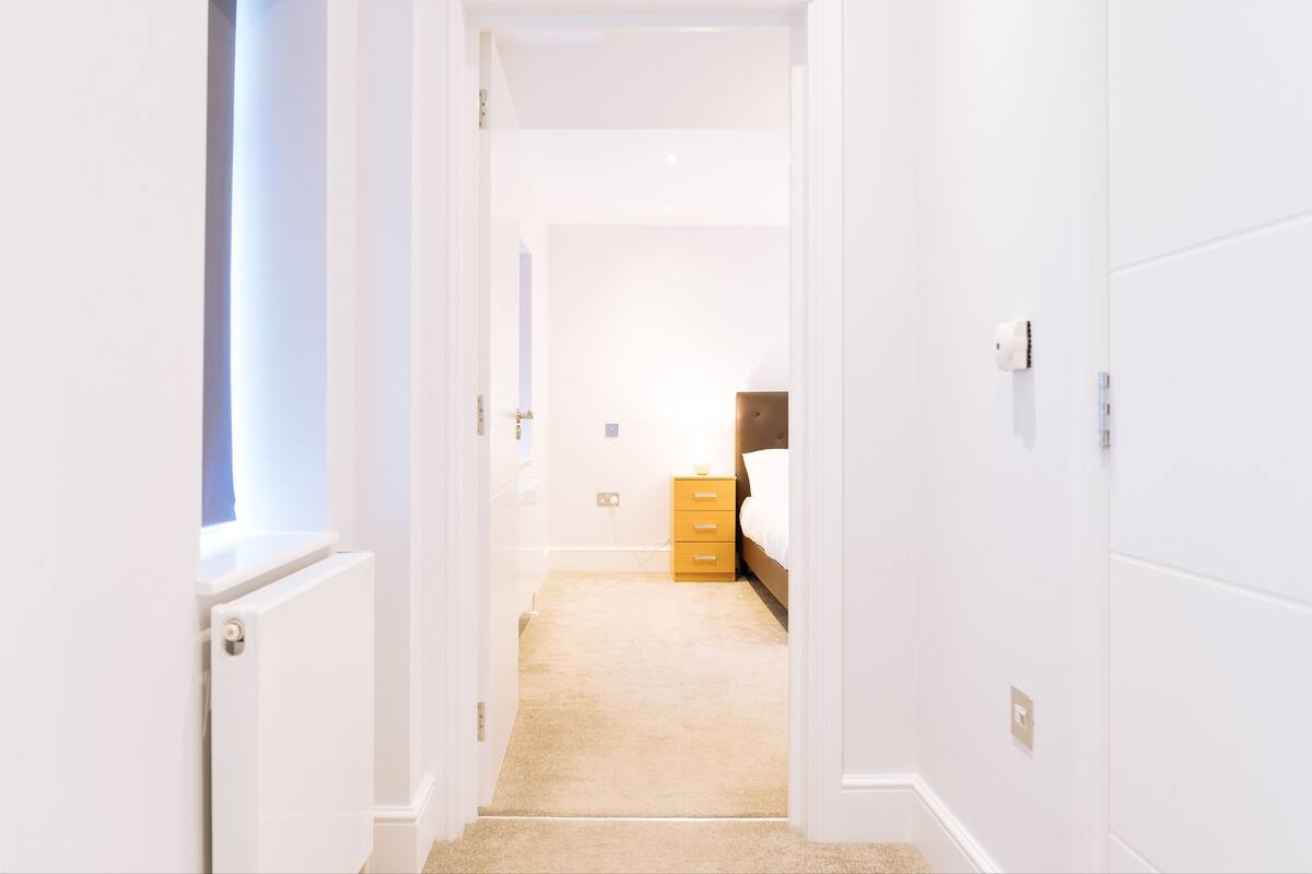 A narrow hallway with white walls and a carpeted floor leading to a bedroom with a bed and a wooden nightstand.