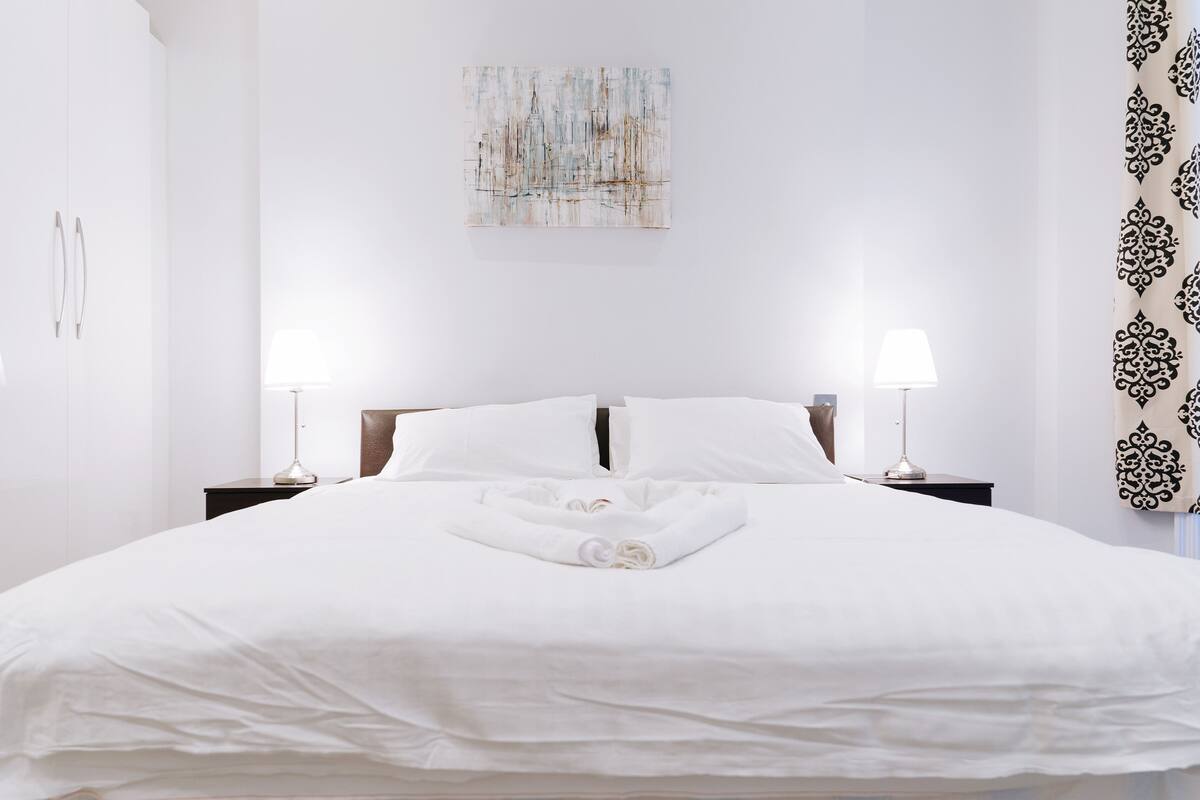A neatly made bed with white linens and a rolled towel in the center, flanked by two bedside tables with lamps, in a minimalist room with a plain wall and a decorative piece above the bed.