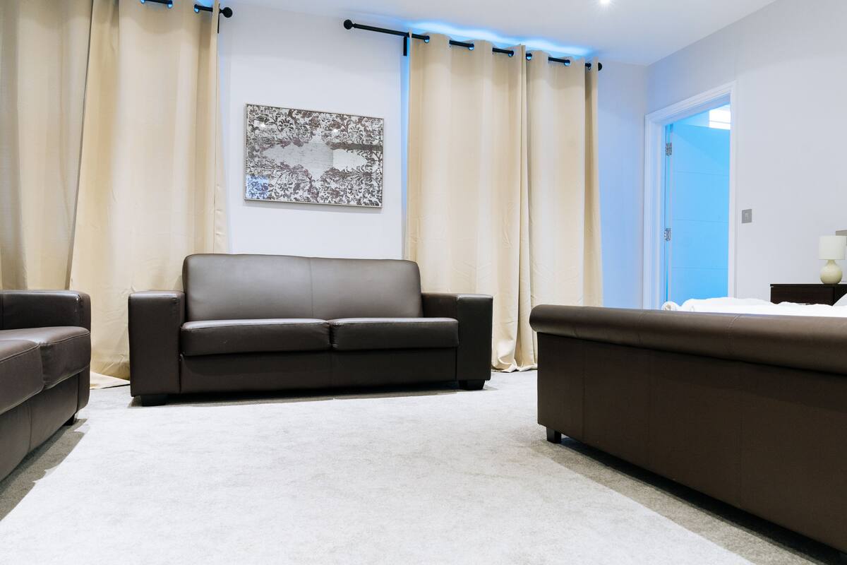 A modern living room with a brown leather sofa, beige curtains, and a framed abstract artwork on the wall.