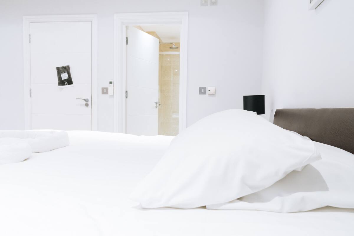 A hotel room with a bed, a nightstand, a wall-mounted lamp, and a door leading to a bathroom.