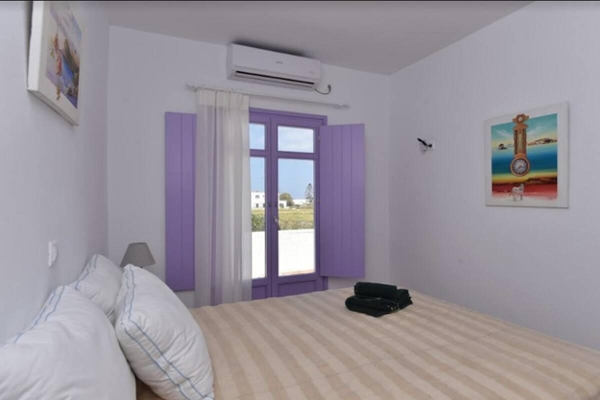 A bedroom with a bed, a window with purple shutters, an air conditioning unit, and a painting on the wall.