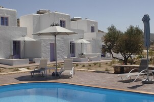 Un área junto a la piscina con edificios blancos, sillas de descanso y sombrillas.