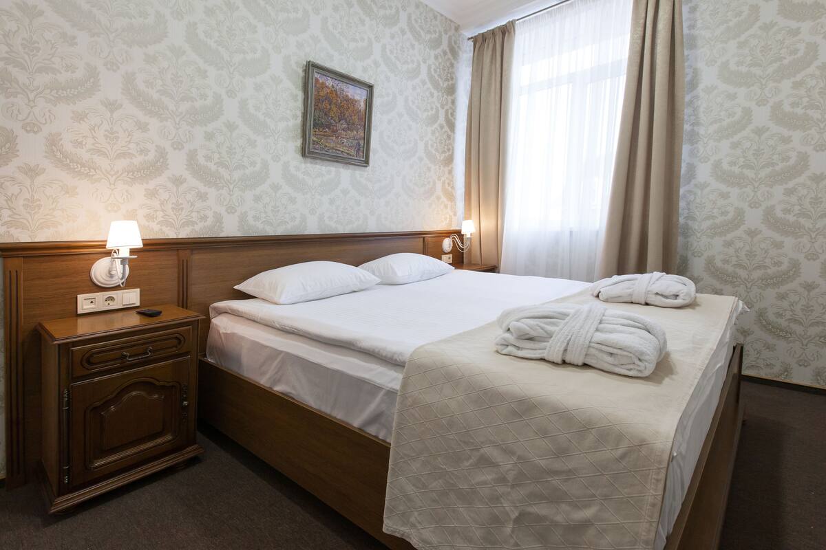A neatly made bed with white linens and a quilted blanket, a wooden nightstand with a lamp, and a painting on the wall.