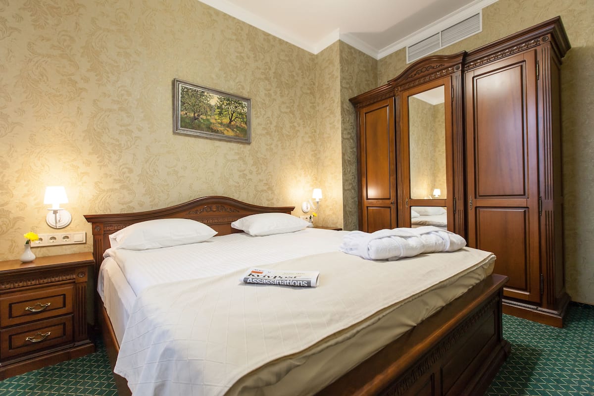 A neatly made bed with white linens, a wooden nightstand with a lamp, a dresser, and a large mirror.