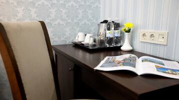 A wooden desk with a magazine, a tray of water bottles, a white vase with yellow flowers, and a white coffee maker.
