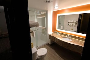 A hotel bathroom with a glass shower enclosure, a sink with a granite countertop, and a mirror with an orange accent wall. - Clarion Inn & Suites Bowling Green (Bowling Green)