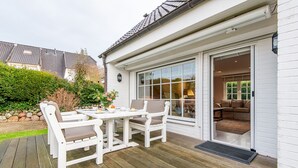 A deck with a table and chairs, a sliding glass door leading to an interior room with a sofa, and a white exterior wall. - Ferienwohnung Rosen/horstweg 5 (Sylt)