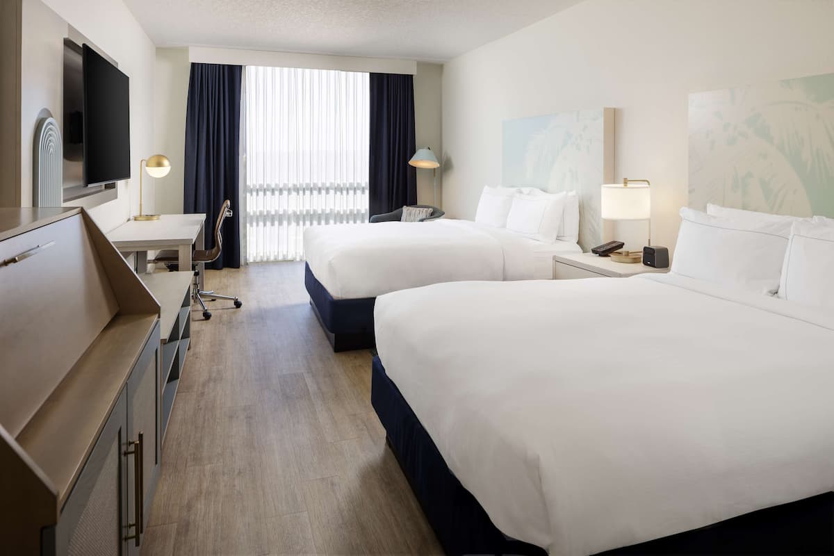 Ein Hotelzimmer mit zwei Betten, einem Schreibtisch, einem Fernseher und einem großen Fenster mit Vorhängen.