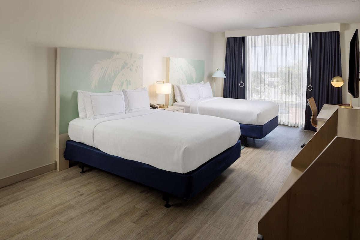 Ein Hotelzimmer mit zwei Betten, einem Schreibtisch, einem Sessel, einem Fernseher und einem großen Fenster mit Vorhängen.