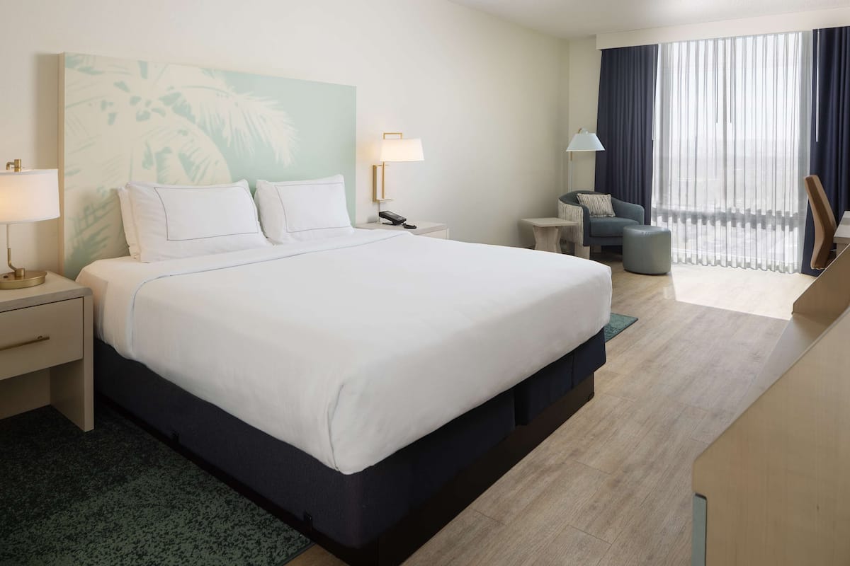 Ein Hotelzimmer mit einem großen Bett, Nachttischen, einem Schreibtisch, einem Stuhl und Blick ins Freie durch leichte Vorhänge.