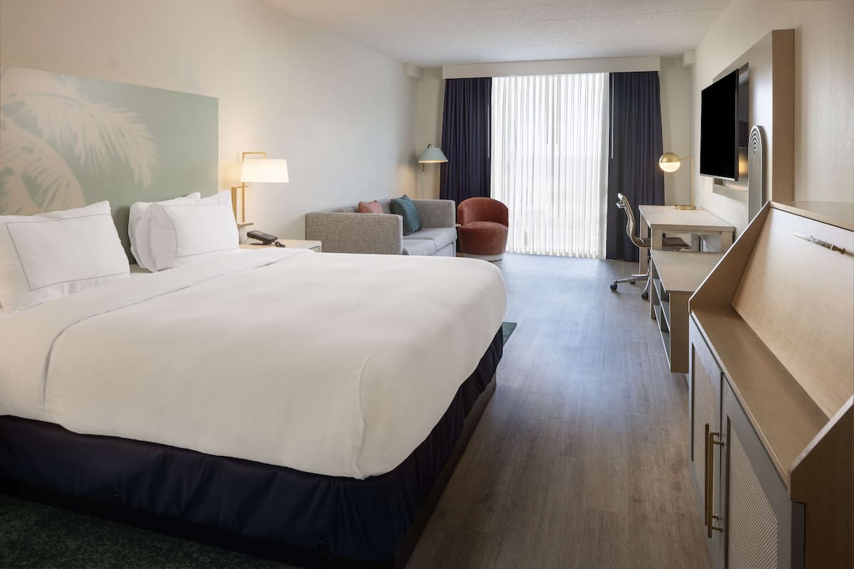 Ein Hotelzimmer mit einem großen Bett, einem Schreibtisch, einem Sessel, einem Fernseher und einem Fenster mit Vorhängen.